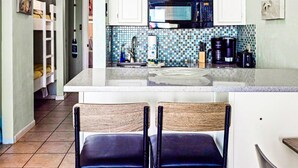 A compact kitchen with white cabinets, a microwave, and a coffee maker. There is a small island with a sink and a chair. - Charming Waterfront Condo (Galveston)