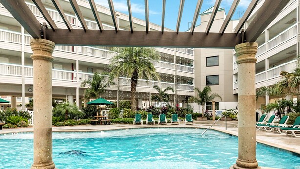 A pool area with a clear blue pool, surrounded by lounge chairs and palm trees, under a covered area with columns. - Charming Ocean View Condo (Galveston)