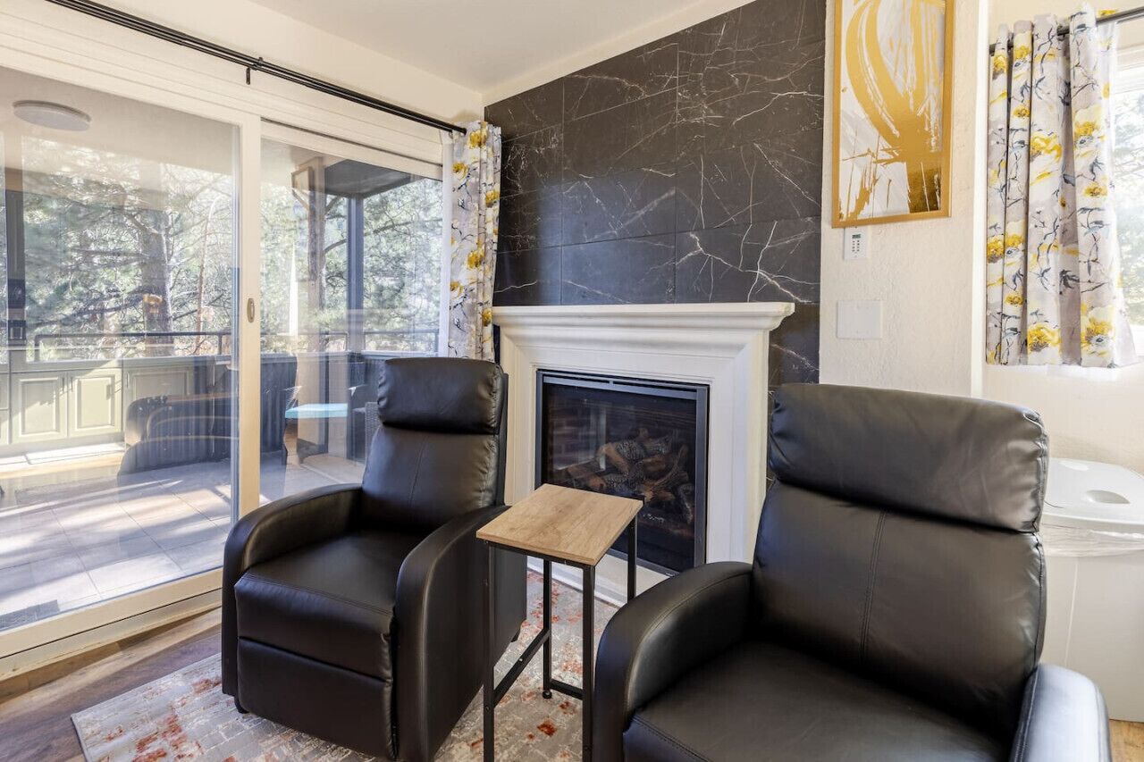 Een moderne woonkamer met een open haard, leren fauteuils, een kleine houten tafel en een glazen deur die leidt naar een terras.