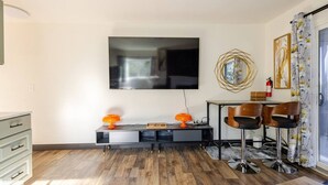 Un salon moderne doté d’un téléviseur à écran plat, d’une table basse, de deux lampes oranges, d’un coin repas avec une table et des chaises, et d’un miroir décoratif accroché au mur.