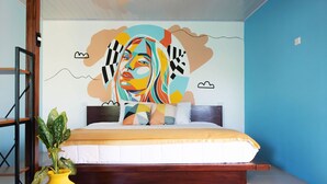 Een slaapkamer met een bed, een gele vaas met een plant, een blauwe muur en een muurschildering.
