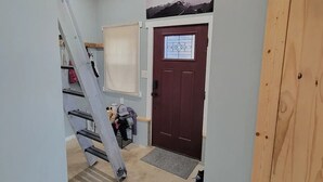 Ein schmaler Korridor mit Holzboden und einer roten Tür mit Fenster. Eine Metallleiter lehnt an der Wand, und ein kleiner Teppich liegt auf dem Boden.