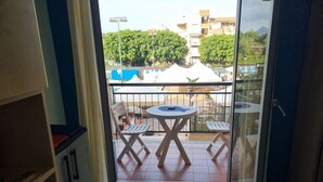 Un balcón con una mesa redonda y dos sillas, piso de baldosas, y vista a edificios y árboles al fondo.