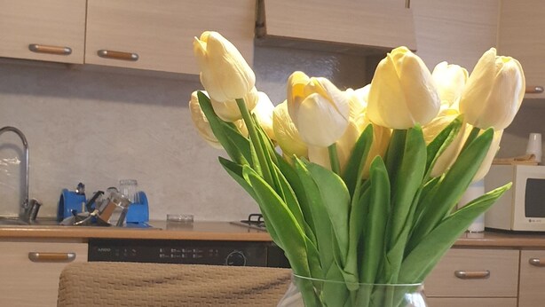A kitchen with wooden cabinets, a dishwasher, and a vase with yellow tulips on a table with a floral tablecloth.
