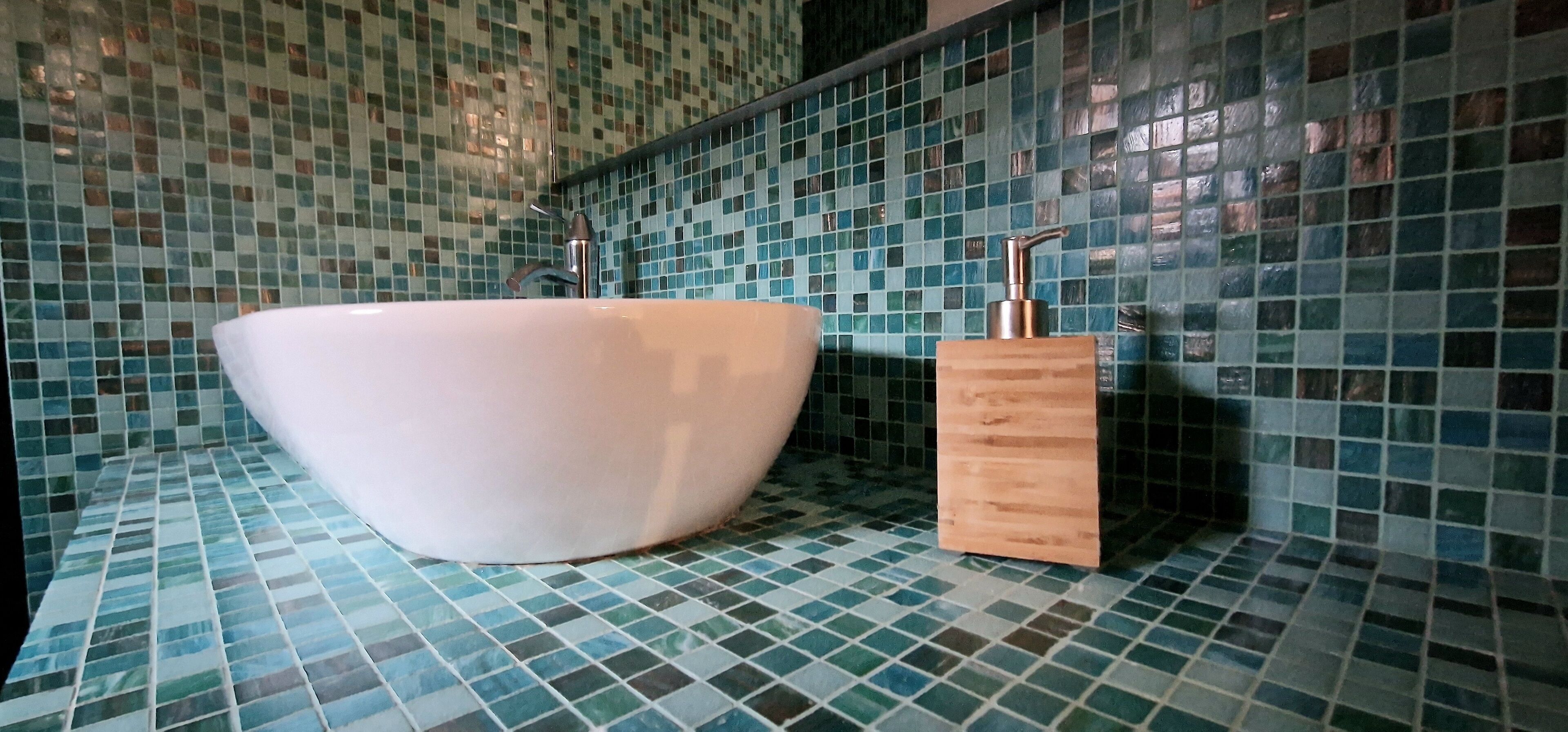 A bathroom with a large white basin and a wooden soap dispenser against a tiled wall.