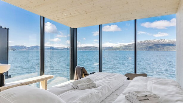 A bedroom with a large window overlooking a lake and mountains. There is a bed with white linens and a folded towel.