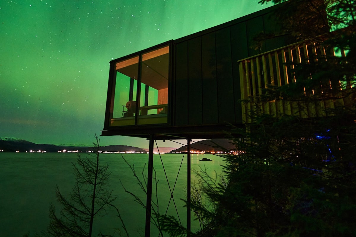 A glass-enclosed cabin with a view of the northern lights over a body of water.
