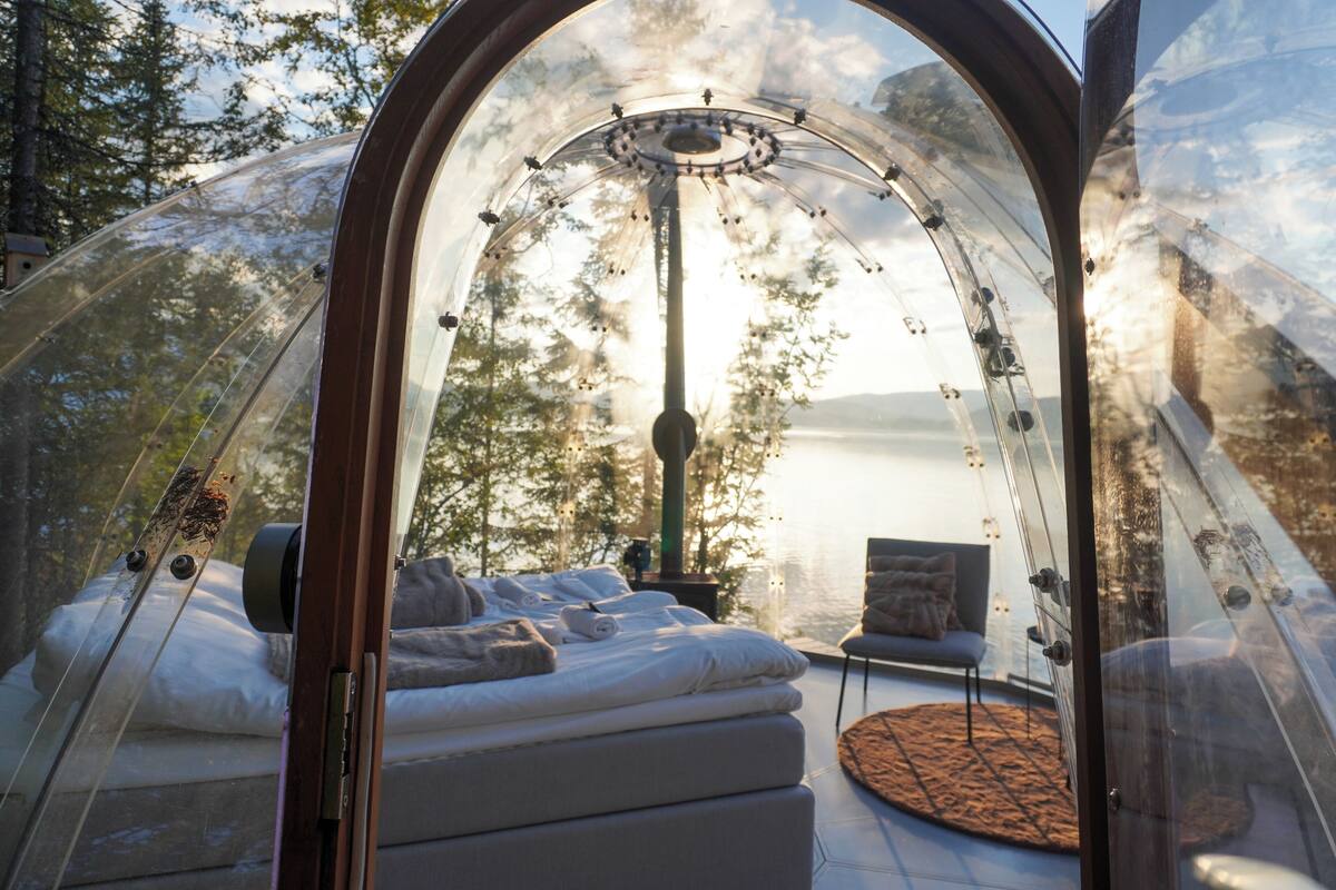A transparent dome structure with a bed and seating area, surrounded by trees and a view of a body of water.