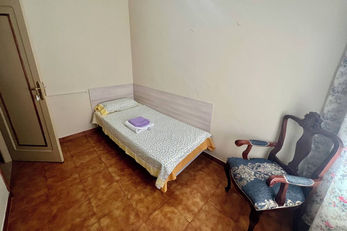 Une petite chambre avec un lit simple, un plancher en bois, une chaise à motifs floraux et une porte.