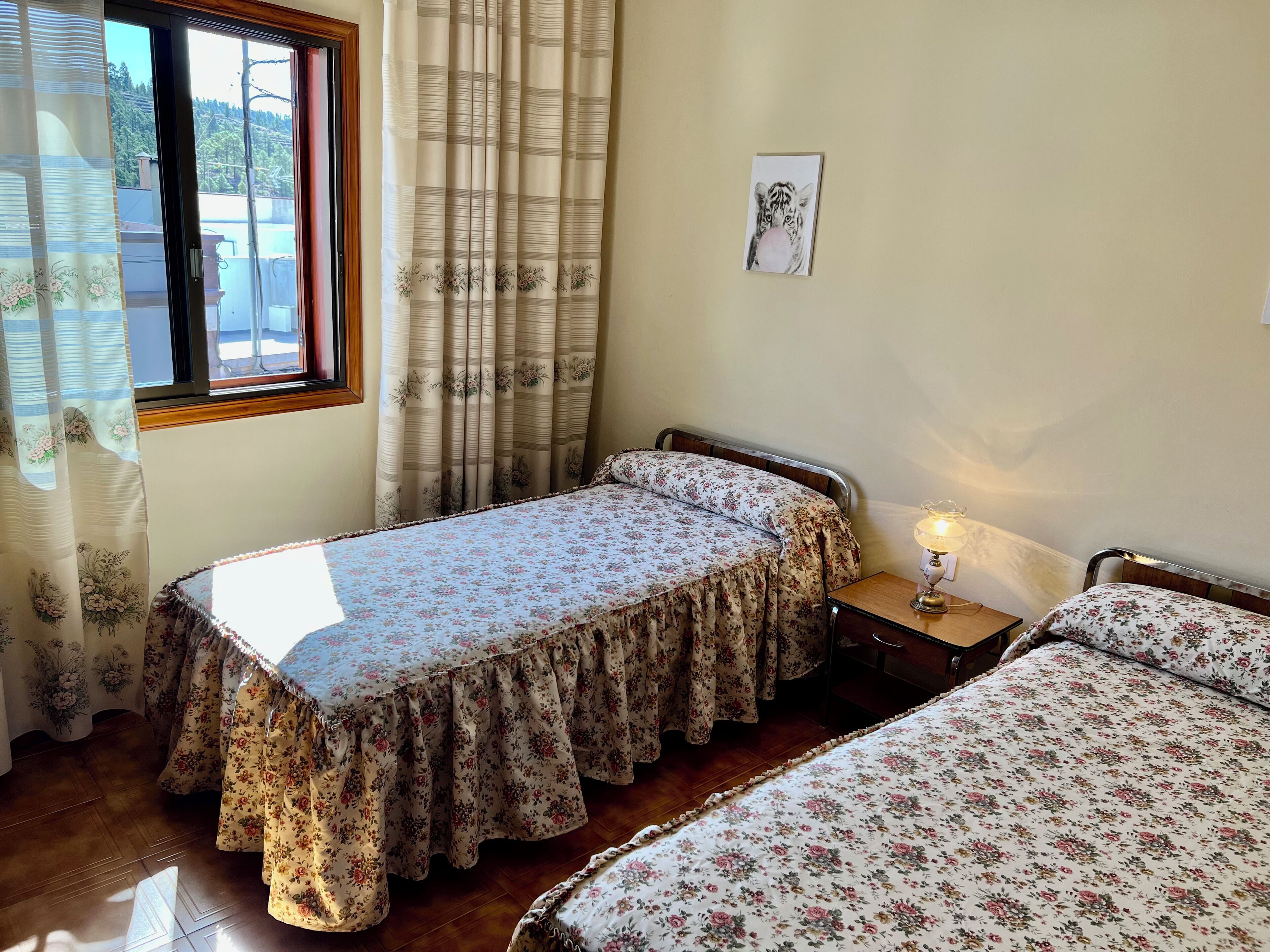 A bedroom with two single beds, a wooden floor, a window with curtains, a bedside table with a lamp, and a framed picture on the wall.