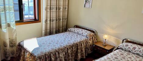 A bedroom with two single beds, a wooden floor, a window with curtains, a bedside table with a lamp, and a framed picture on the wall.