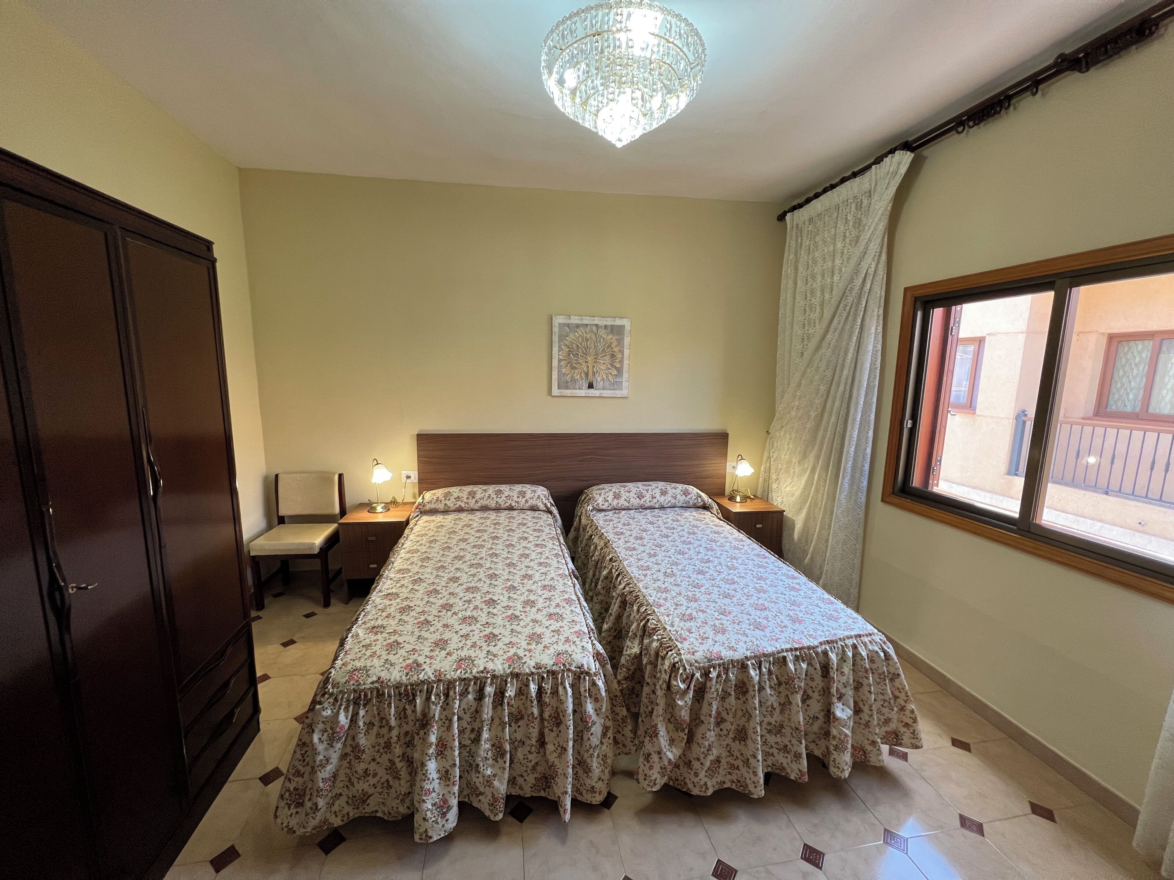 Quarto de hotel com duas camas, guarda-roupa de madeira, escrivaninha com cadeira, mesa de cabeceira com luminárias, candelabro, janela com vista e um quadro na parede.