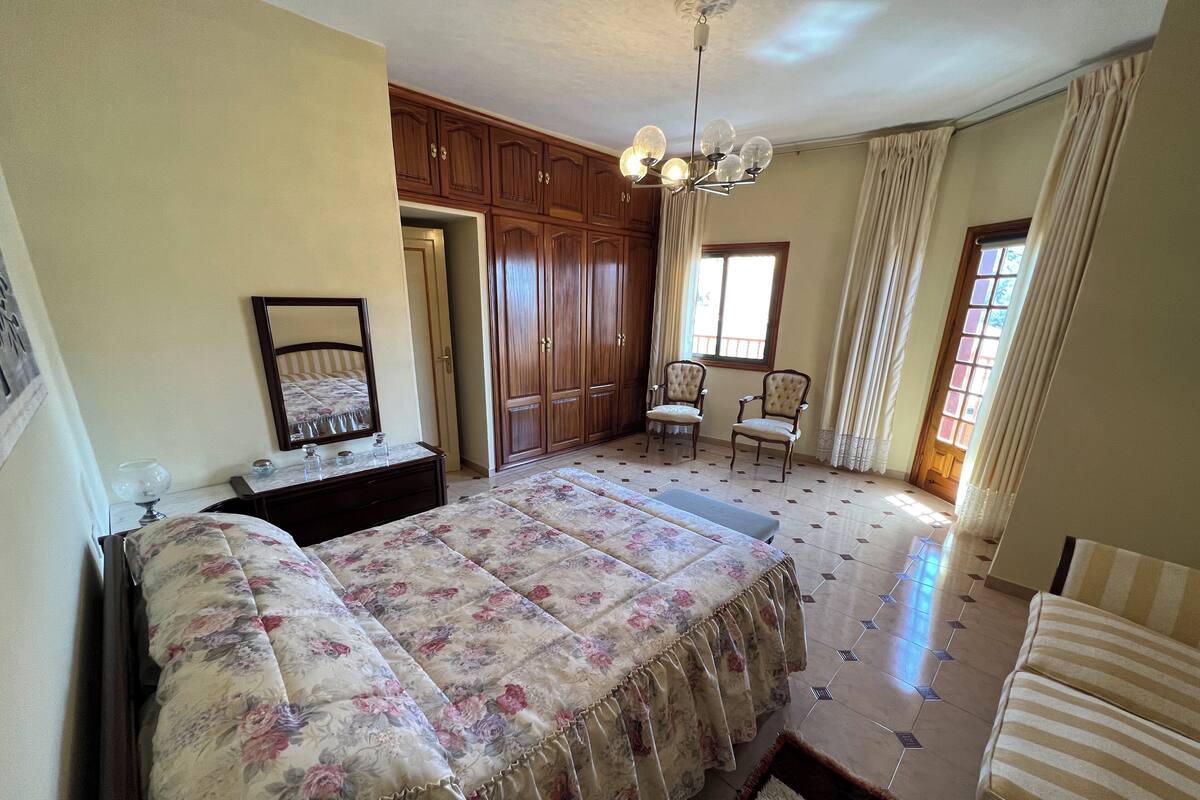 Une chambre à coucher avec un grand lit, une commode, un miroir, deux chaises, une armoire et une fenêtre avec des rideaux.
