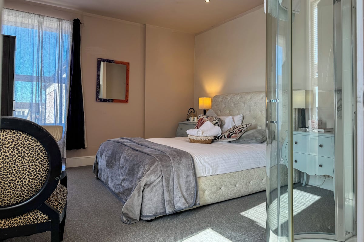 A bedroom with a bed, a bedside table with a lamp, a chair, a glass shower enclosure, and a window with curtains.