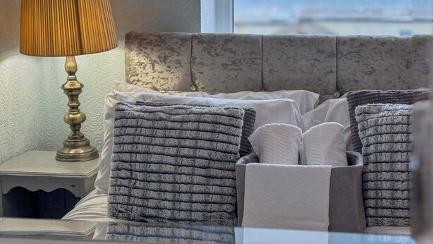 A bed with a grey headboard, a bedside table with a lamp, and a view of the sea through a window. - The Chatwal Boutique Hotel (Blackpool)