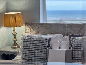 A bed with a grey headboard, a bedside table with a lamp, and a view of the sea through a window.