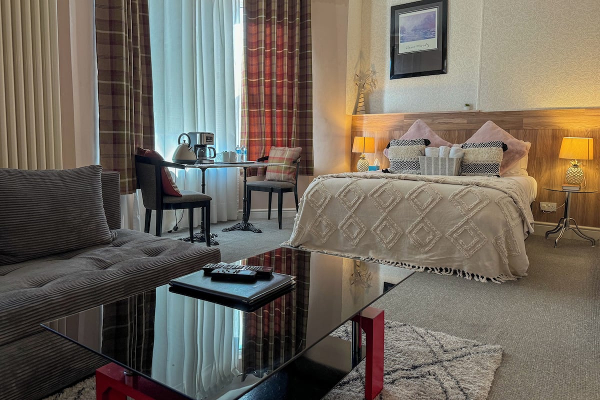A hotel room with a sofa, a glass coffee table, a bed with patterned bedding, a bedside table with a lamp, and a framed picture on the wall.