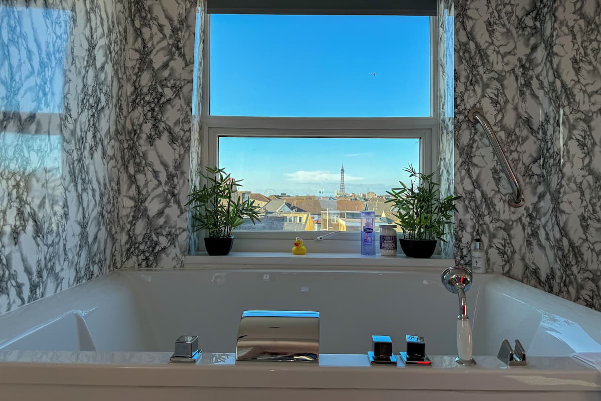 A bathroom with a large window, two potted plants, a bathtub, and a handrail.