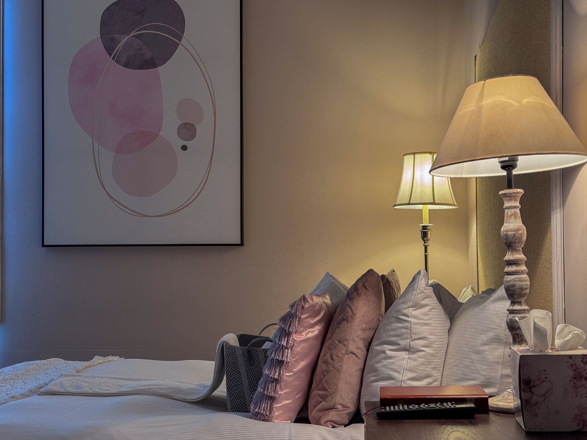 A neatly made bed with a white comforter and several pillows, a bedside table with a lamp and remote control, and a framed abstract artwork on the wall.