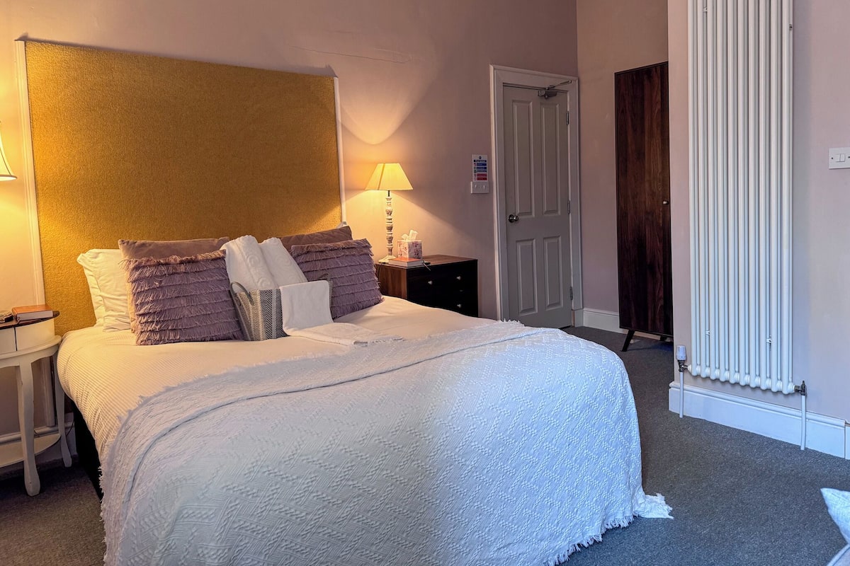 A neatly made bed with a white comforter and multiple pillows, a bedside lamp, and a nightstand with a book.
