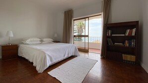 Un dormitorio con cama, mesitas de noche, una estantería, una ventana con cortinas y vistas al mar.