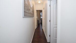 Ein schmaler Flur mit dunklem Holzboden, weißen Wänden und einer Tür rechts. Ein gerahmtes Bild hängt an der linken Wand.
