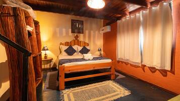 Een houten bed met kussens, een nachtkastje met een lamp en een groot raam met gordijnen.