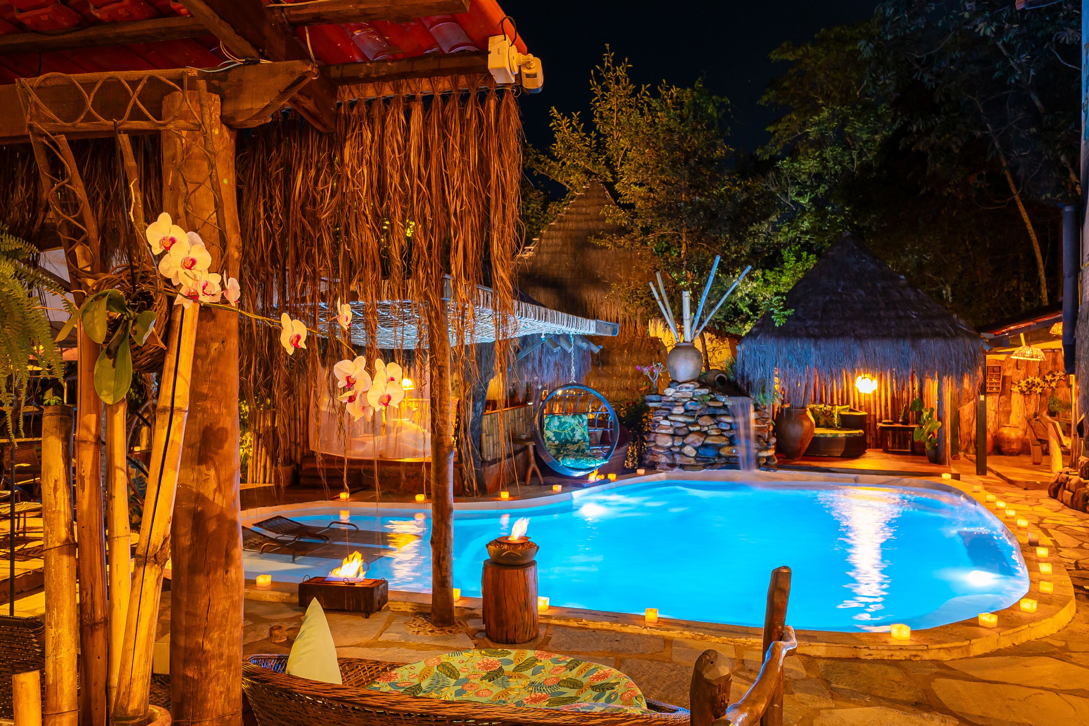 A pool area with a thatched-roof structure, a hanging chair, and a small waterfall feature.