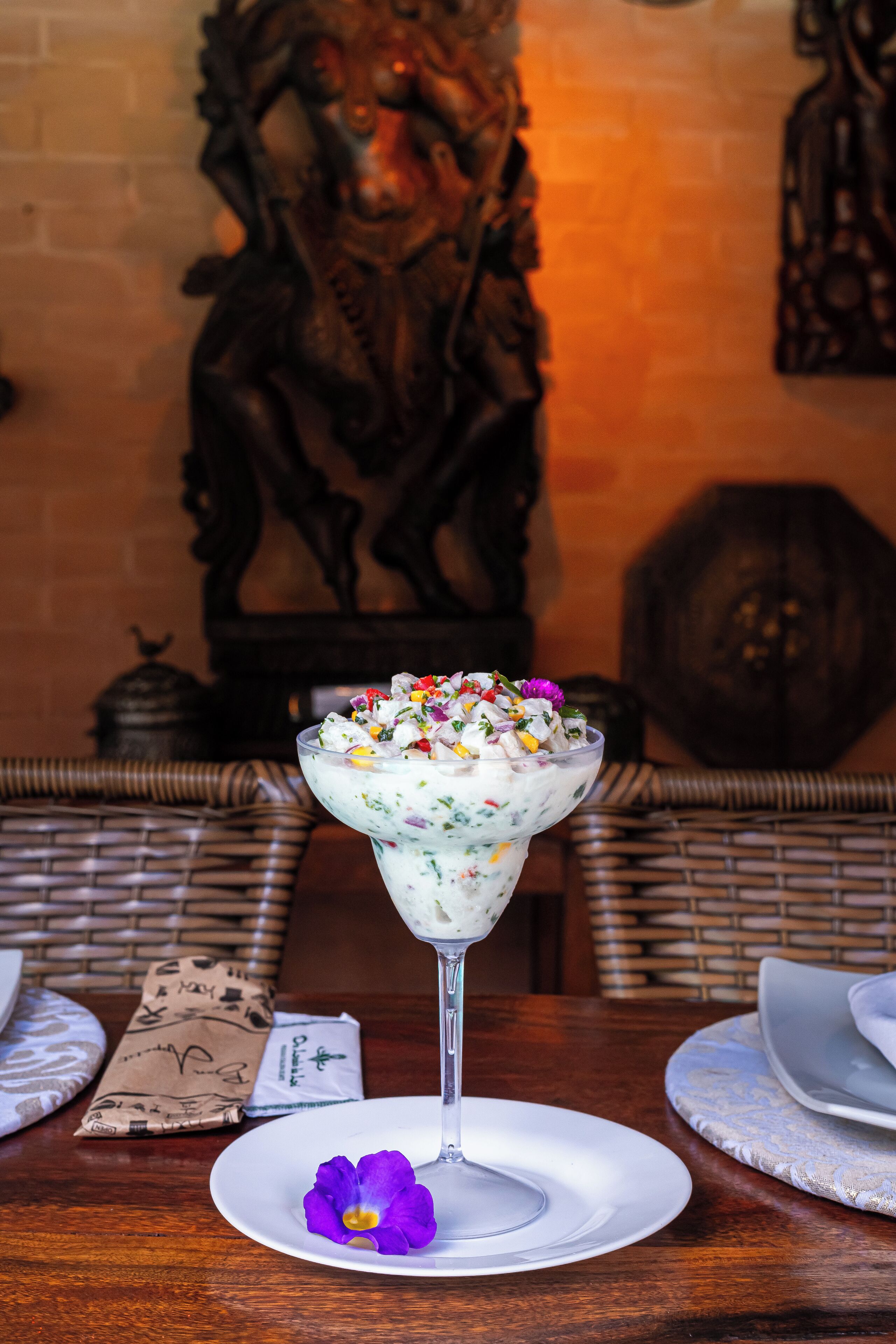 A dessert in a glass on a table with a wicker chair and a decorative wall in the background.