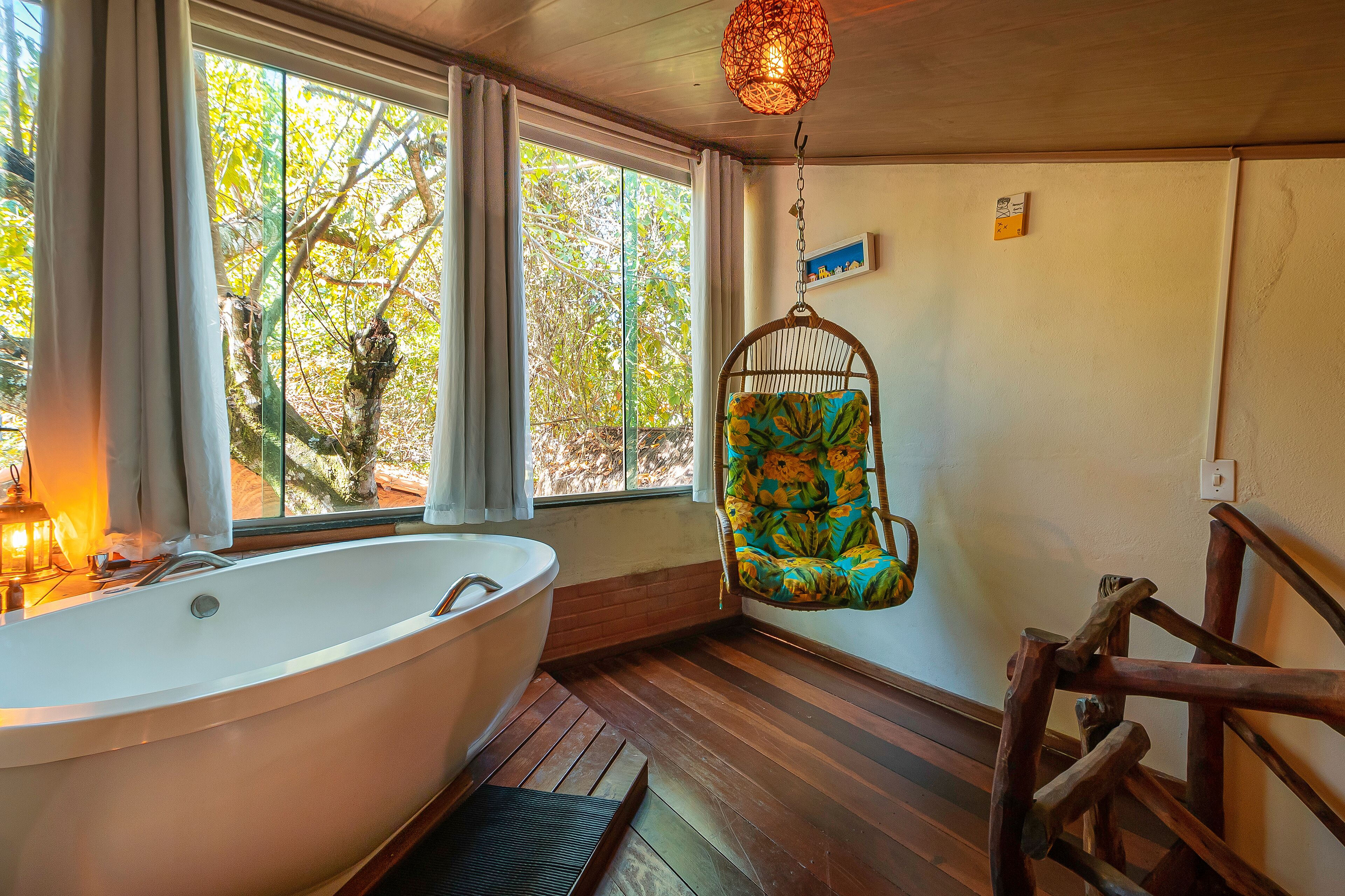 A bathroom with a freestanding bathtub, wooden flooring, a hanging chair, and a view of trees through large windows.