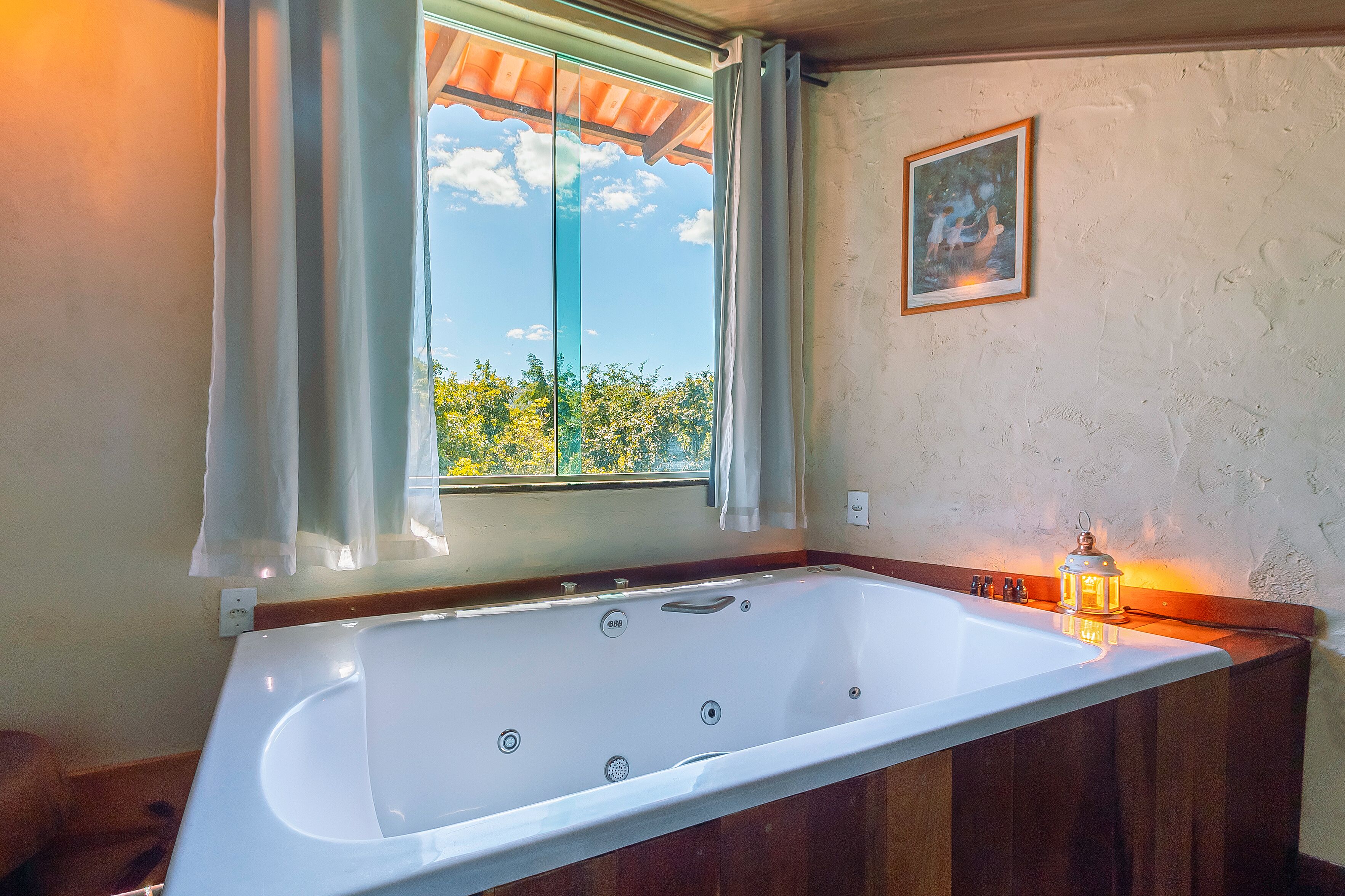 A large bathtub with a view of trees through a window, a framed picture on the wall, and a lit lantern on the side.