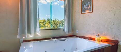 A large bathtub with a view of trees through a window, a framed picture on the wall, and a lit lantern on the side.