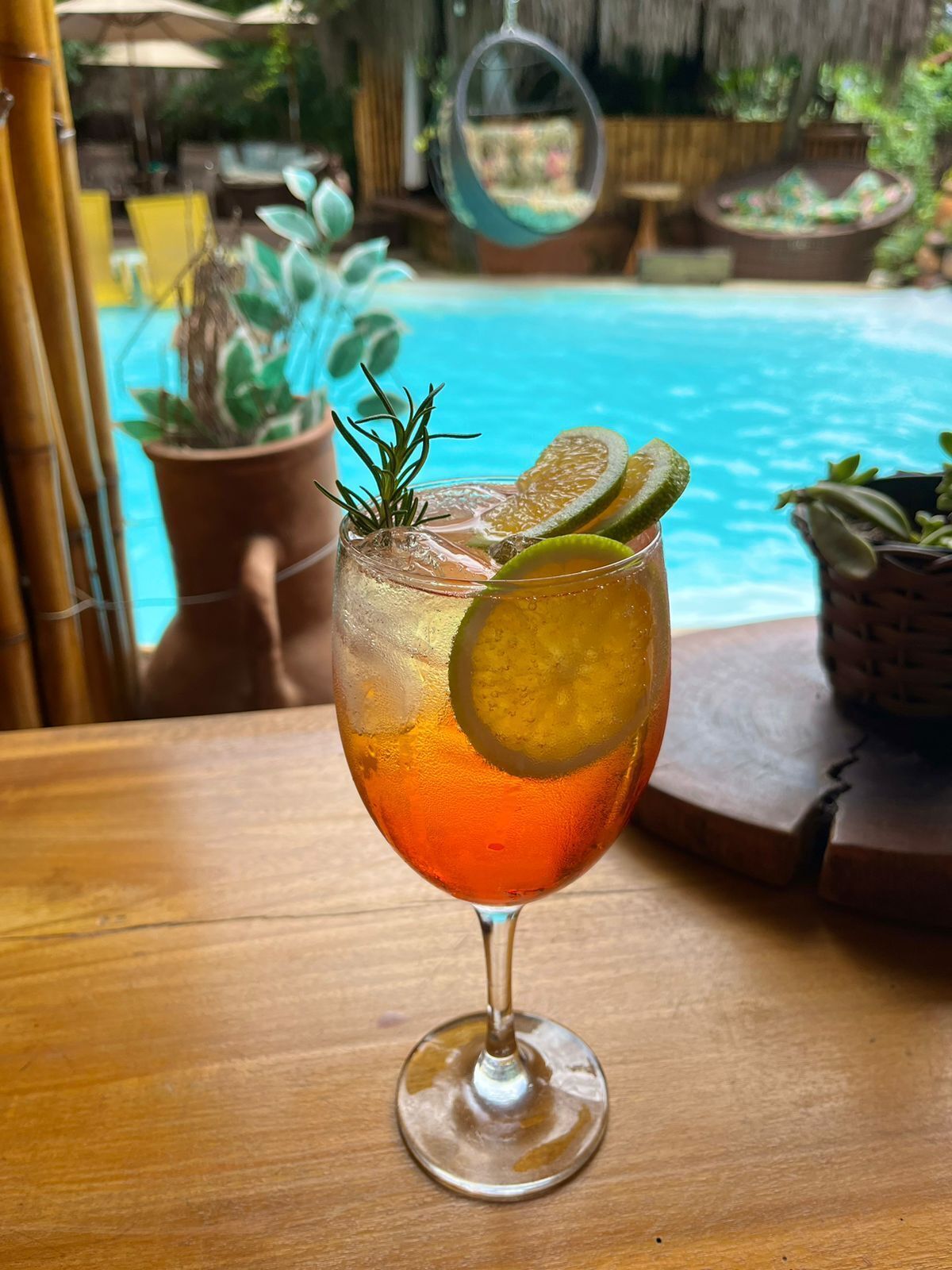 Un vaso de cóctel con una rodaja de lima y ramita de romero sobre una mesa de madera junto a una piscina.