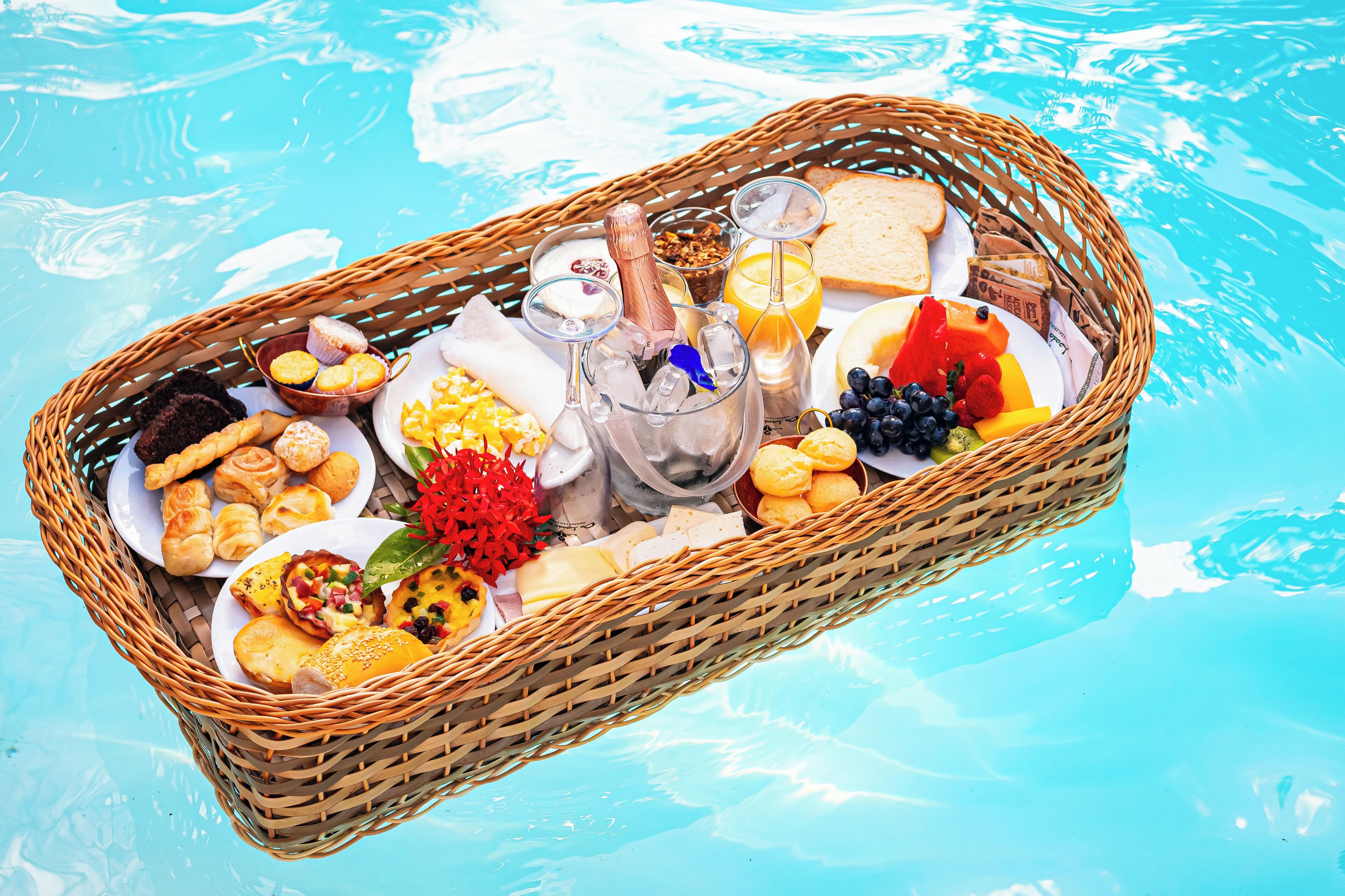 Una cesta de mimbre llena de diversos productos para el desayuno, colocada al lado de una piscina.