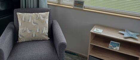 A grey armchair with a seagull pillow, a wooden shelf with a starfish and a book, and a window with blinds.