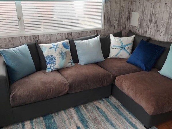 A sectional sofa with multiple cushions, a striped rug, and a window with blinds. - Dog Friendly Caravan on the Northumberian Coast (Whitley Bay)