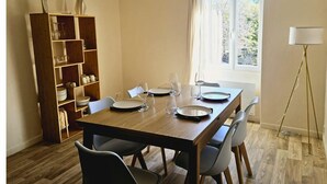 A dining area with a wooden table set for four, surrounded by chairs. A bookshelf with various items is to the left. - Apartment - shaded terrace (Marsanne)