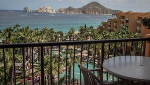A balcony with a view of a pool and palm trees, overlooking the ocean and rocky cliffs.