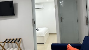 Quarto moderno com cama, TV fixada na parede, sofá azul e porta para outro cômodo.