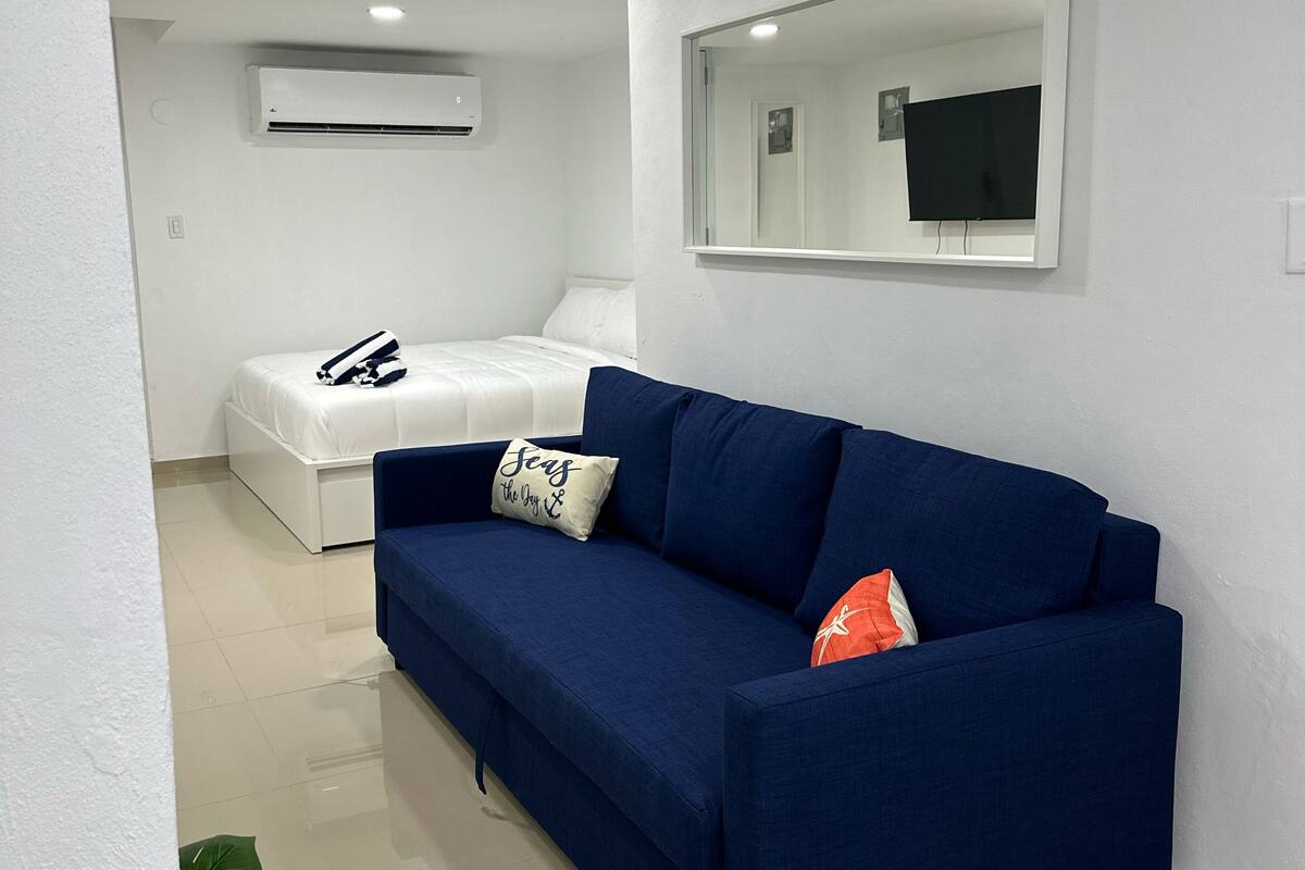 A modern room with a blue sofa, a white bed, a flat-screen TV mounted on the wall, and an air conditioning unit above the bed.