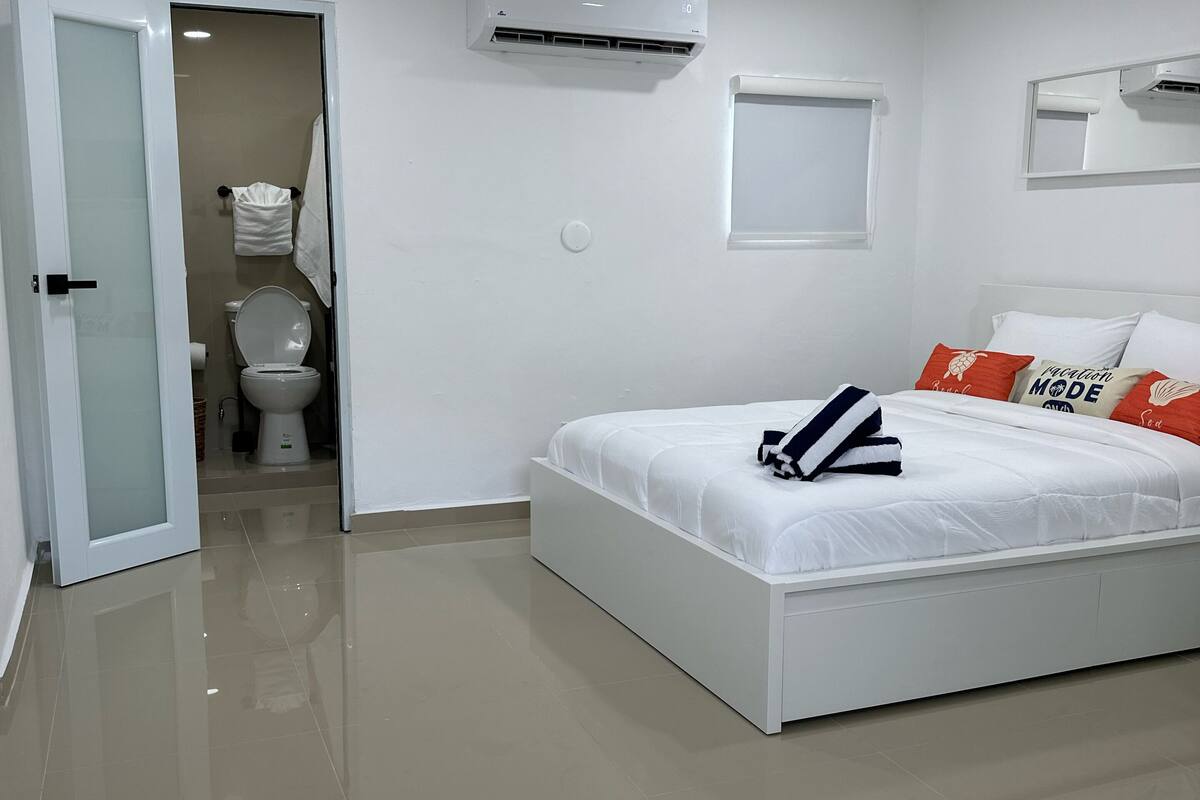 A modern bedroom with a white bed, a bathroom visible through an open door, and a ceiling-mounted air conditioner.