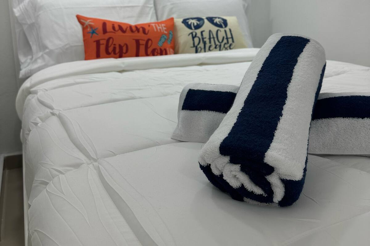 A neatly made bed with a white comforter and a blue and white striped towel. There are three pillows on the bed, each with different text and designs.
