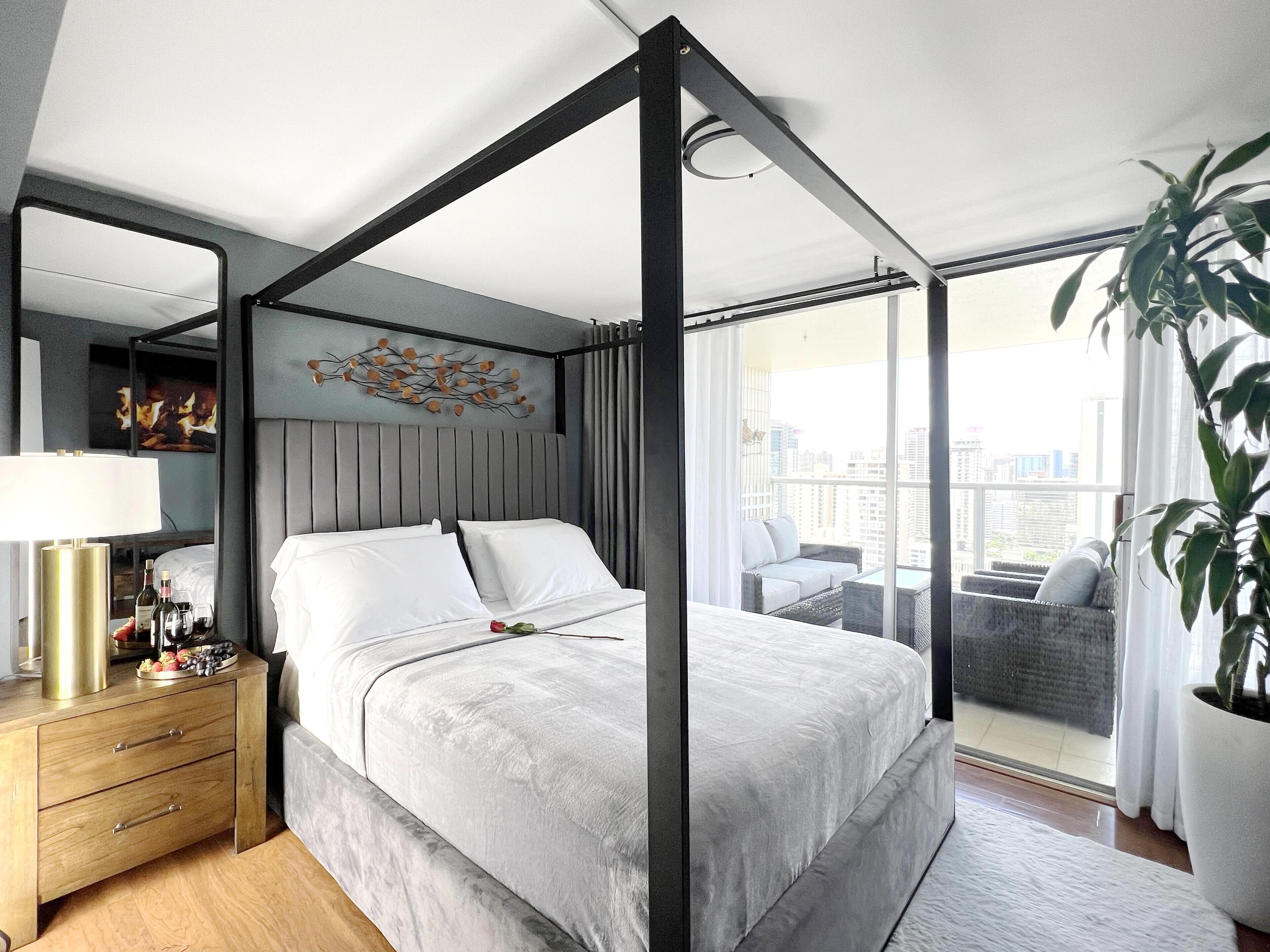 A modern bedroom with a four-poster bed, a bedside table with a lamp, and a view of the city through large windows.