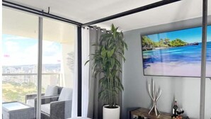 A modern bedroom with a large window, a bed, a TV, a plant, and a balcony with a view of the city.