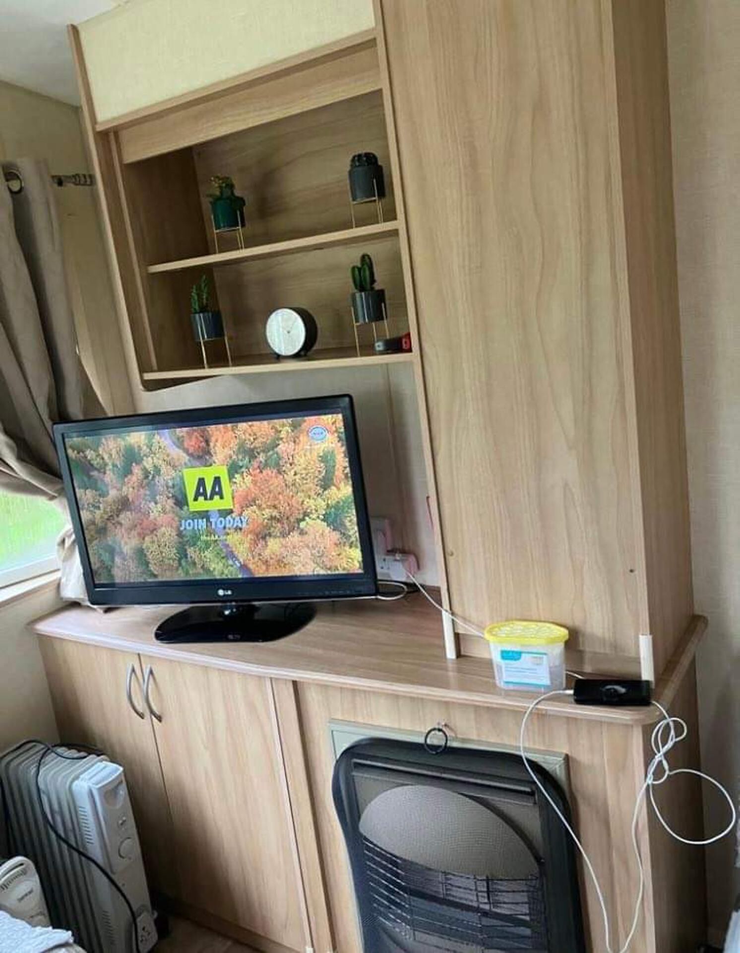Een ruimtelijk RV-interieur met een houten entertainmentmeubel, een flatscreen-tv waarop een advertentie wordt getoond, en een kleine kitchenette.