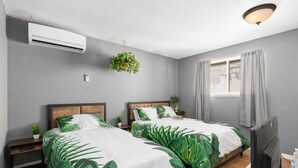 Un dormitorio con dos camas, un ventilador de techo, aire acondicionado, una planta colgante, una mesita de noche, un televisor y una ventana con cortinas.