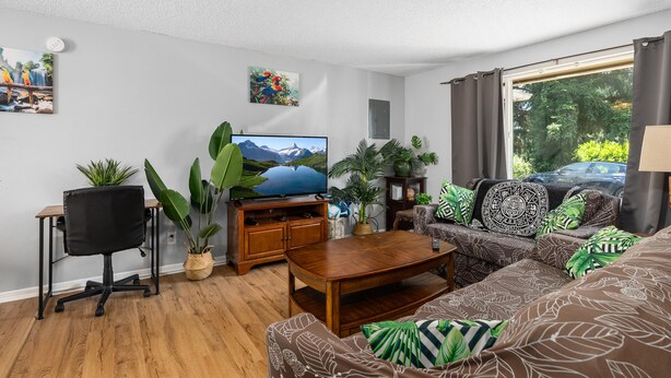 Una sala de estar con una mesa de centro de madera, una silla de oficina negra, un sofá con cojines estampados, un televisor sobre un mueble de madera y varias plantas en macetas.