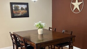 A dining room with a wooden table, chairs, a vase with flowers, a framed picture, a ceiling light, and a wall star decoration. - The Den - Family Friendly Close to Fiesta Texas (San Antonio)