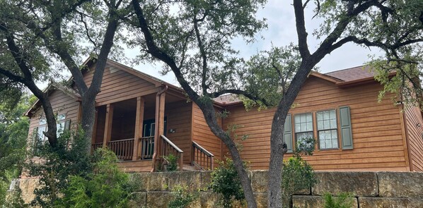 A wooden house with a porch, stone wall, and large trees.
