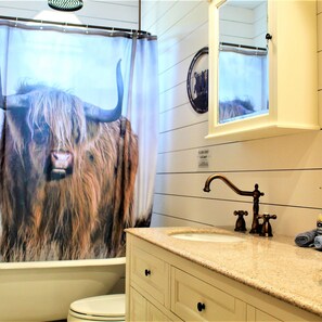 A bathroom with a shower curtain featuring a bull's face, a white toilet, a sink with a vintage faucet, and a mirror above the sink. - The Rustic Inn- Family Friendly Near Fiesta Texas (San Antonio)